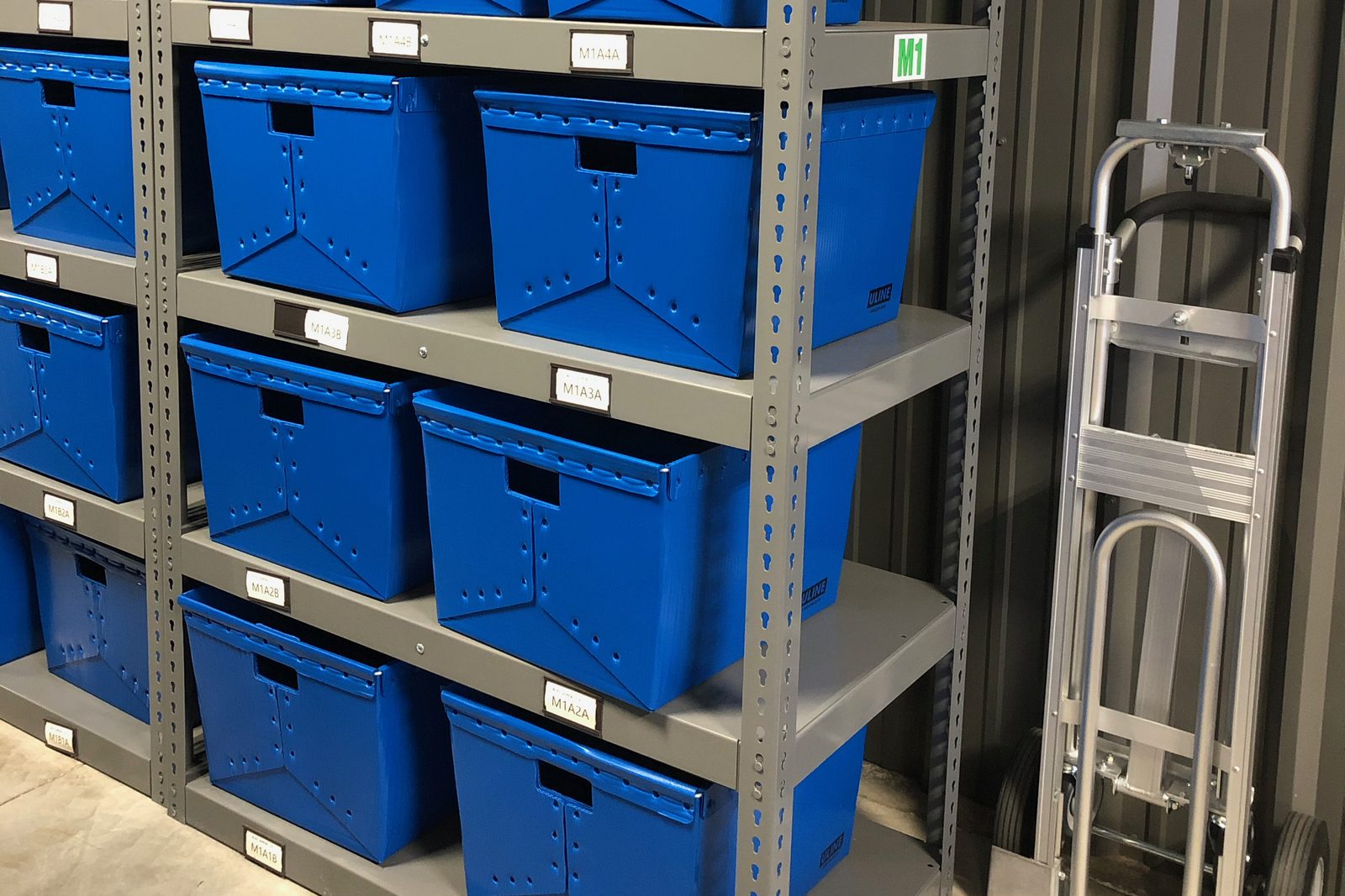 Blue storage totes on labeled steel shelving beside a hand truck